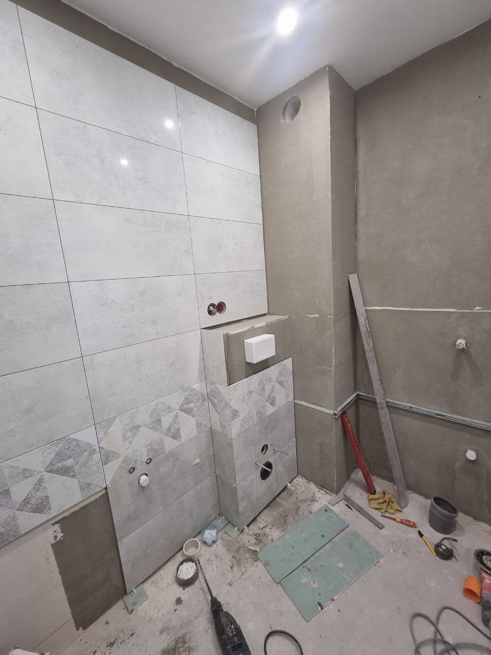 Bathroom wall half-tiled mid-installation, lower board still exposed with tools nearby